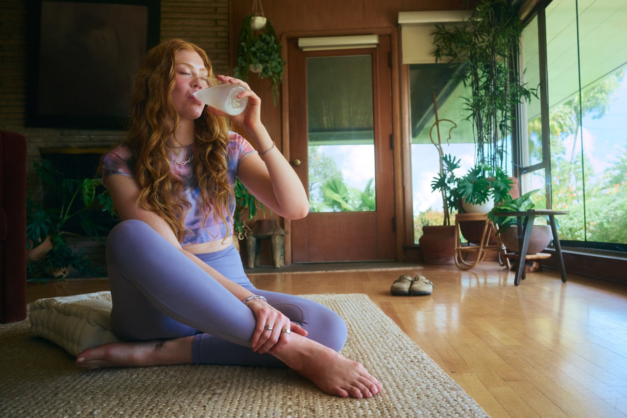 Woman sitting on a mat in a cozy living room, drinking Liquid I.V. for hydration and relaxation